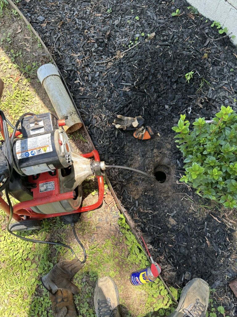 Drain cleaning machine clearing a clogged outdoor sewer cleanout near a home foundation.
